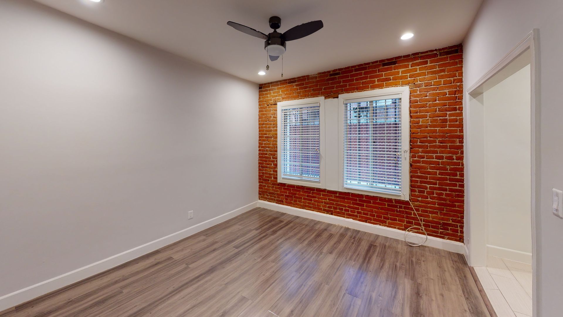 Empty room with brick accent wall, windows, and wood flooring.