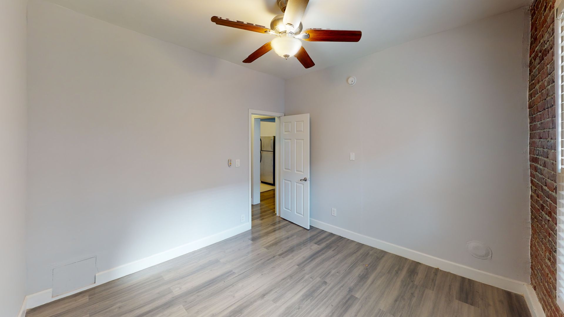 Empty bedroom with light gray walls, wood-look flooring, ceiling fan, and door to a kitchen.