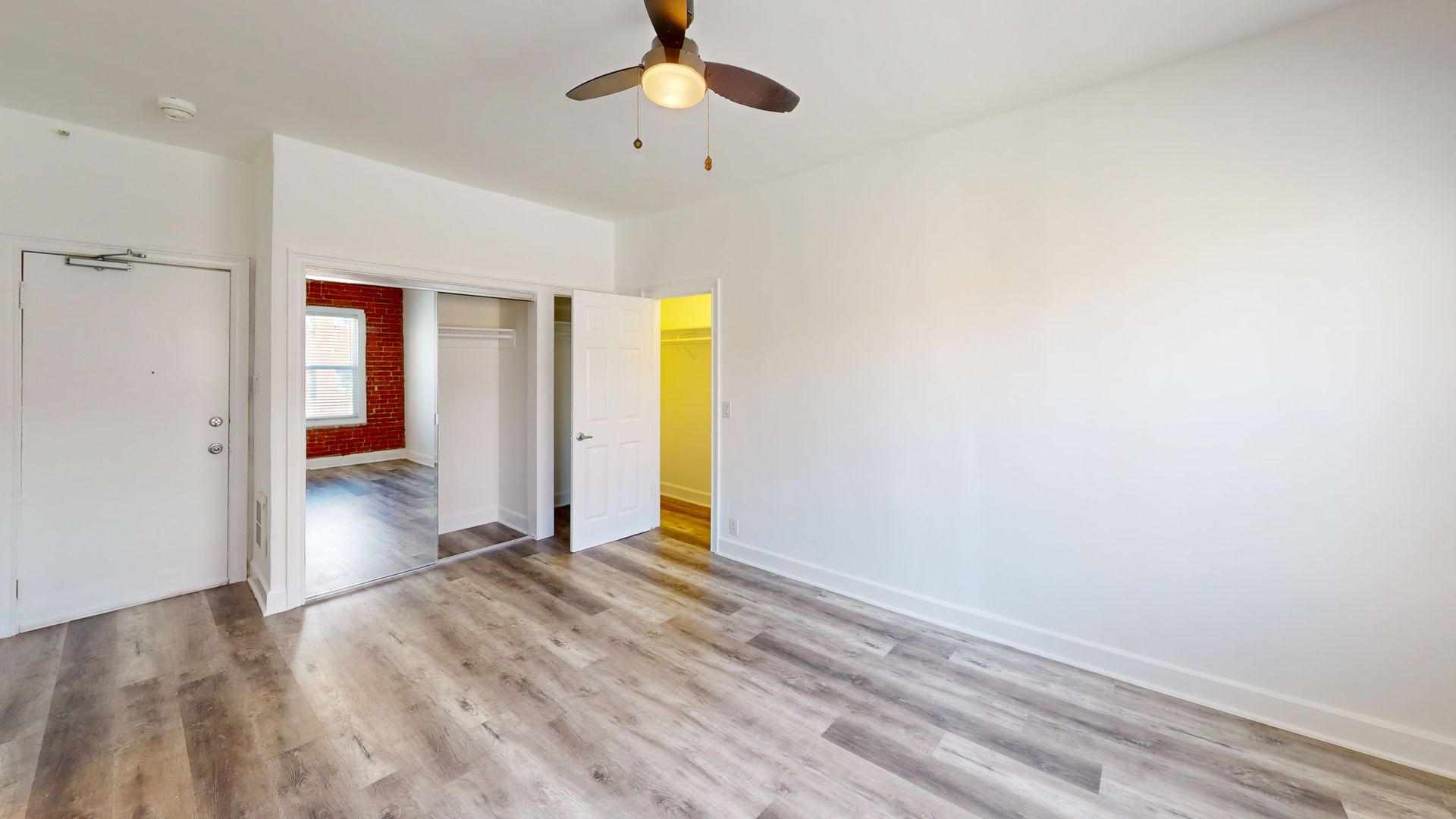 Empty room with wood floors, white walls, closet, doorway to another room with exposed brick.