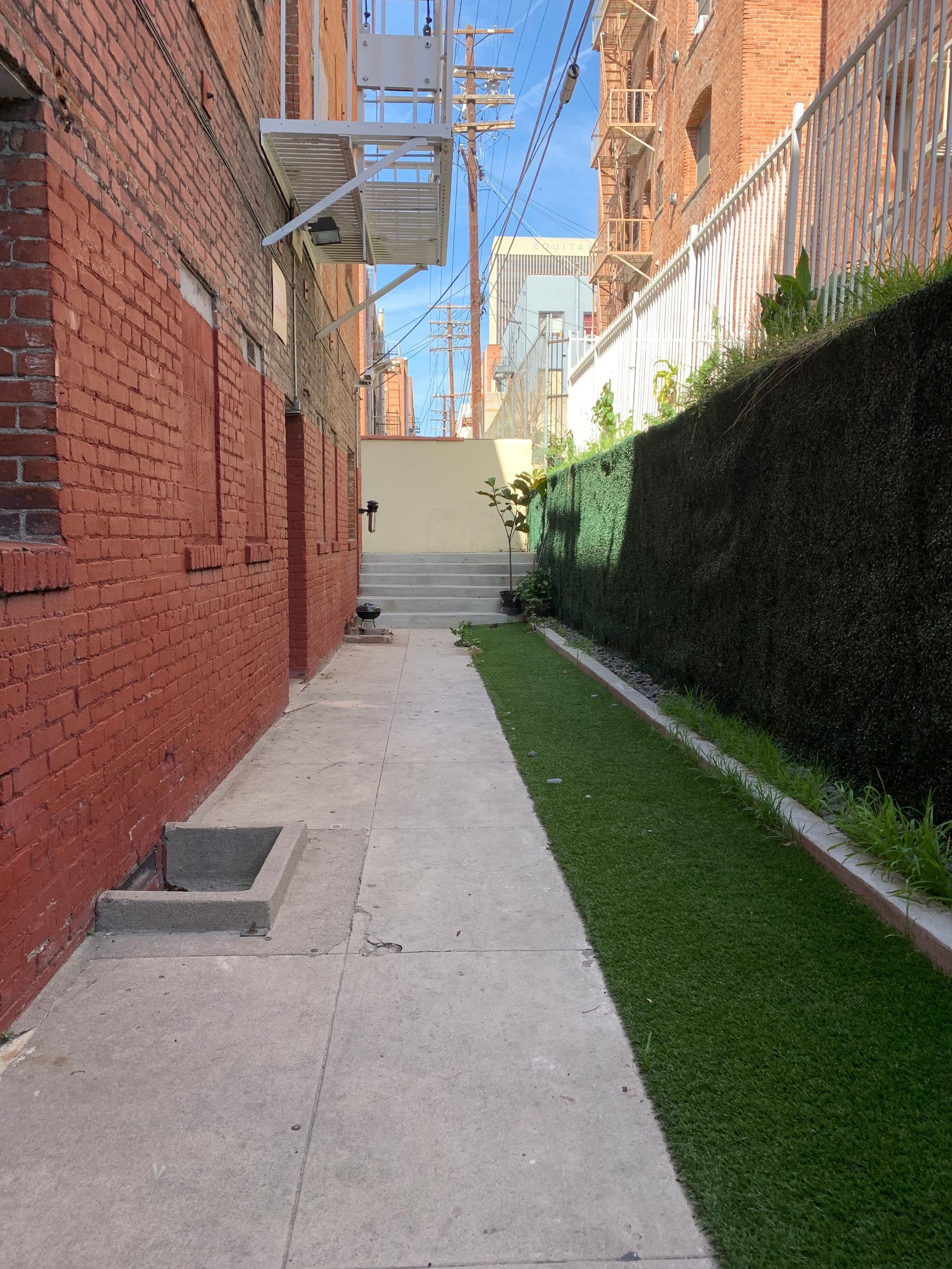 Narrow alley with red brick building, concrete sidewalk, green grass, and stairs leading upwards.