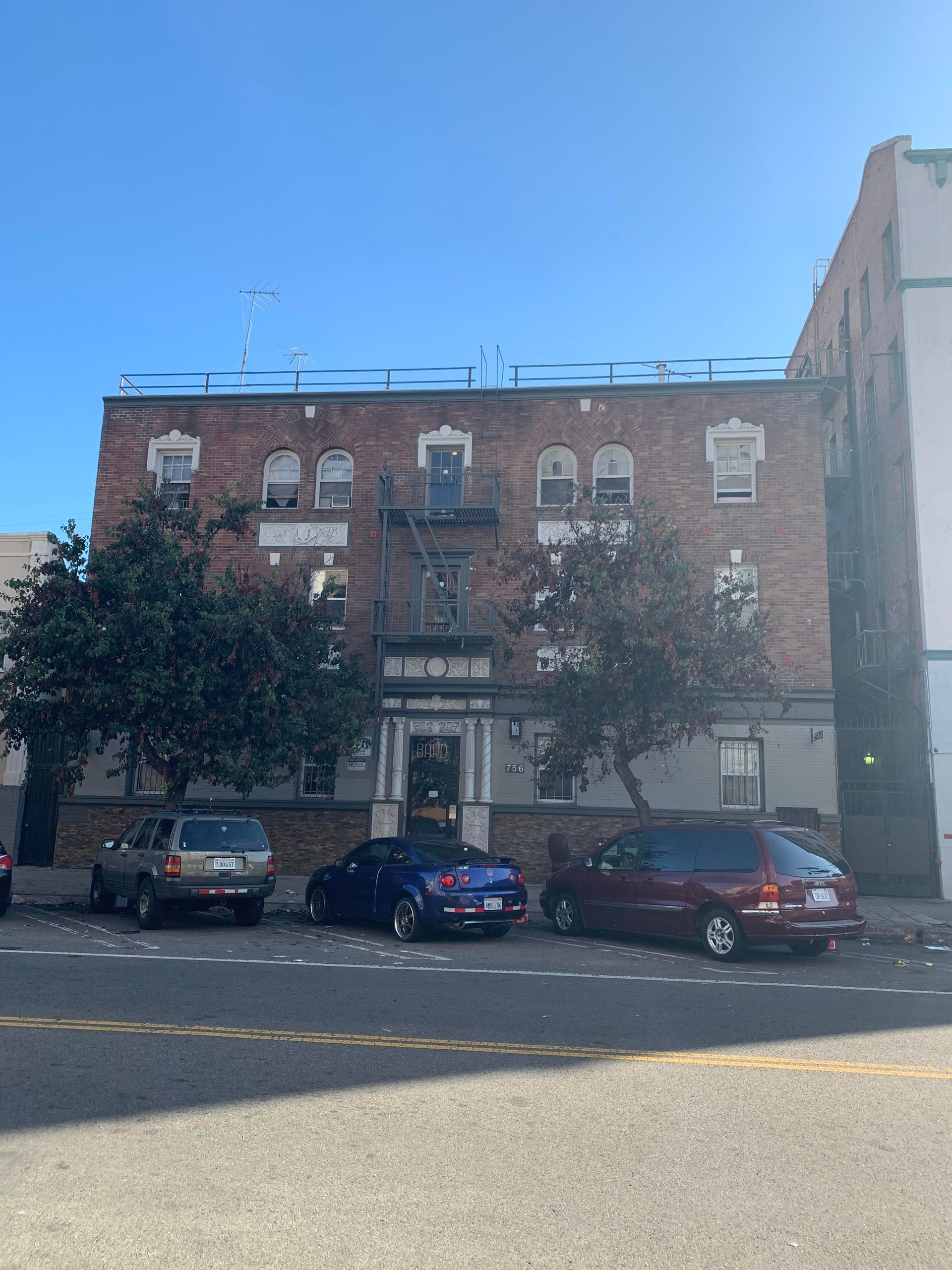 Brick apartment building with parked cars on a sunny street. Fire escape visible.