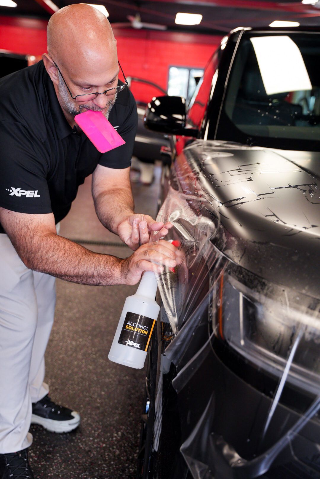 Man applying film to a black car's headlight. He sprays a bottle, holding a pink tool, in a shop.