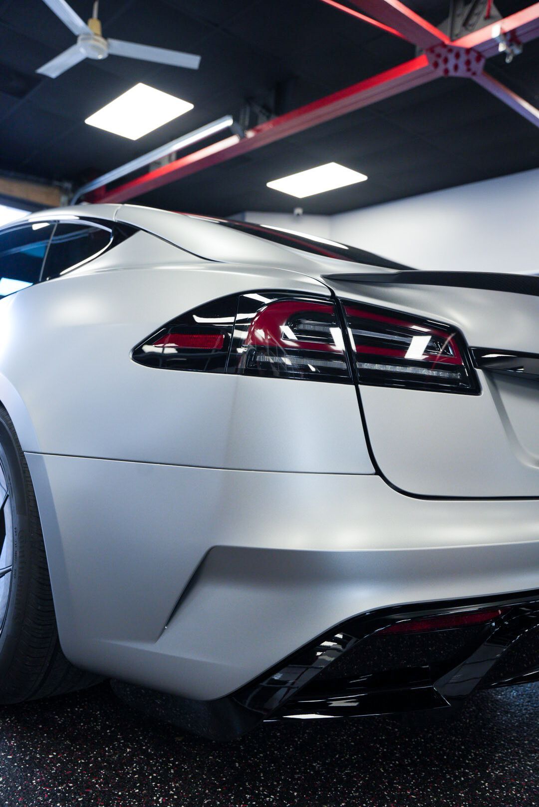 Silver Tesla sedan's rear corner, matte finish. Black spoiler and taillights. Garage interior with red accents.