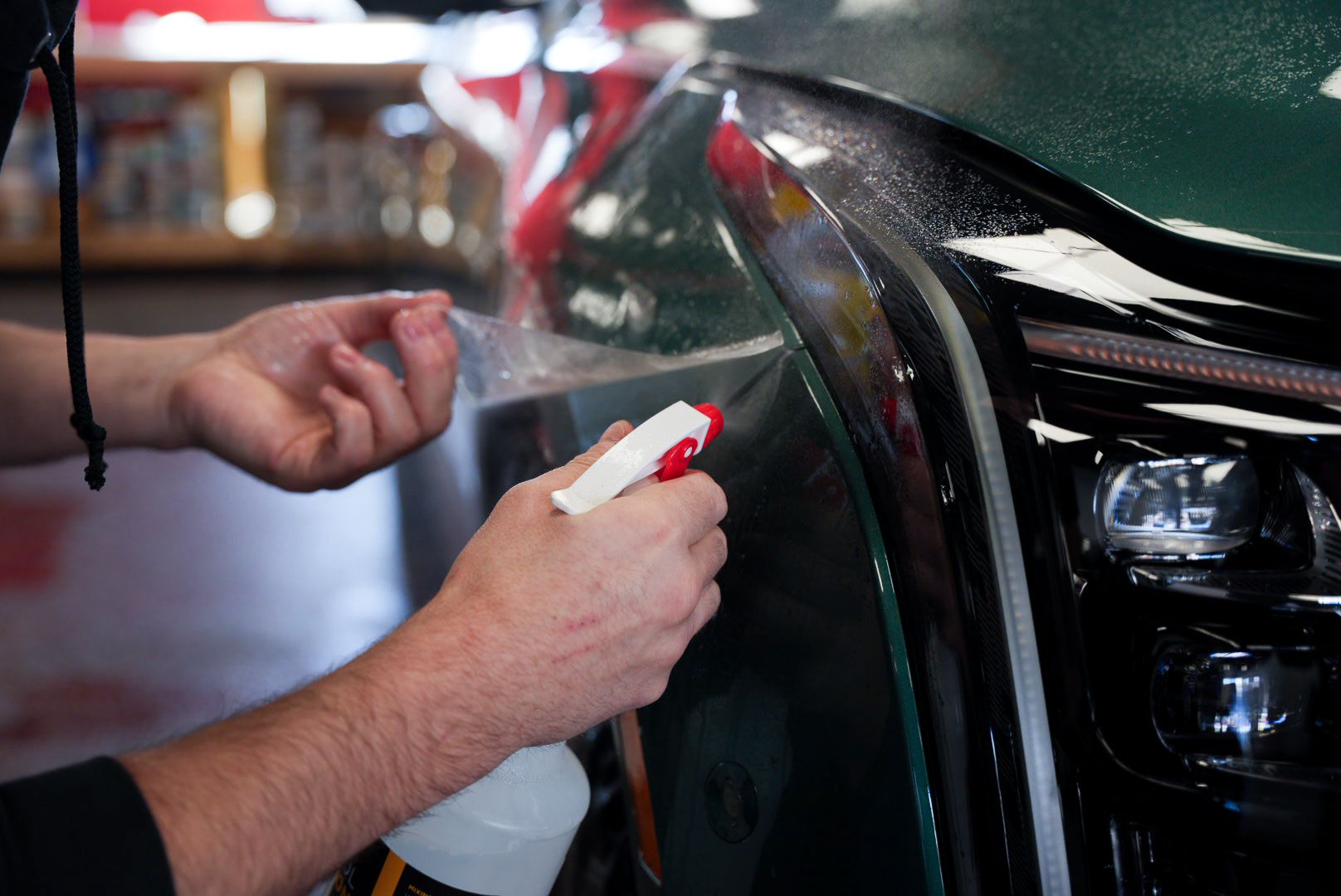 A person sprays liquid on a car's headlight, preparing to apply clear protective film. The car is dark green.
