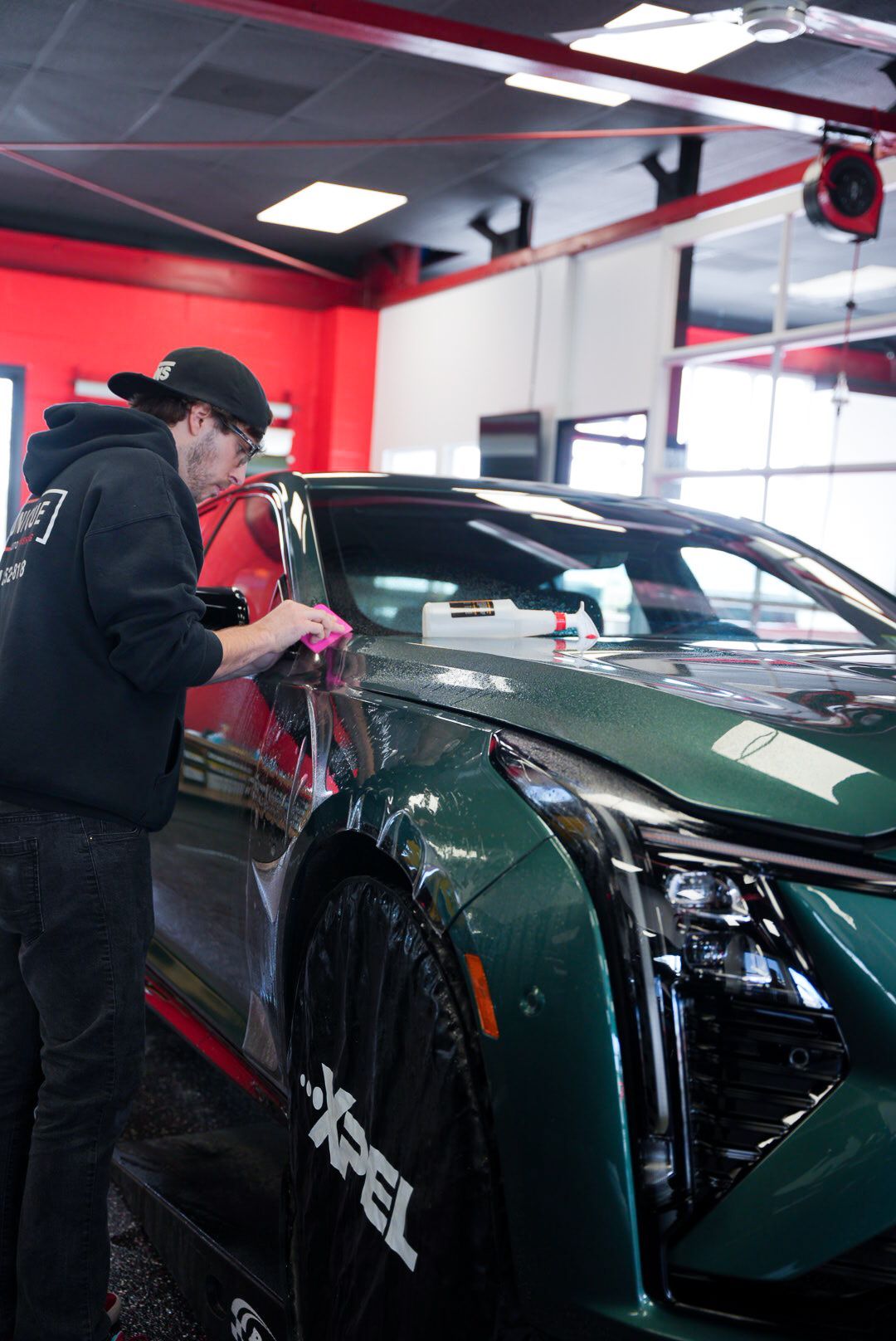 A person in a black hoodie applies film to a green car in a shop. The car has an XPEL logo on the wheel.