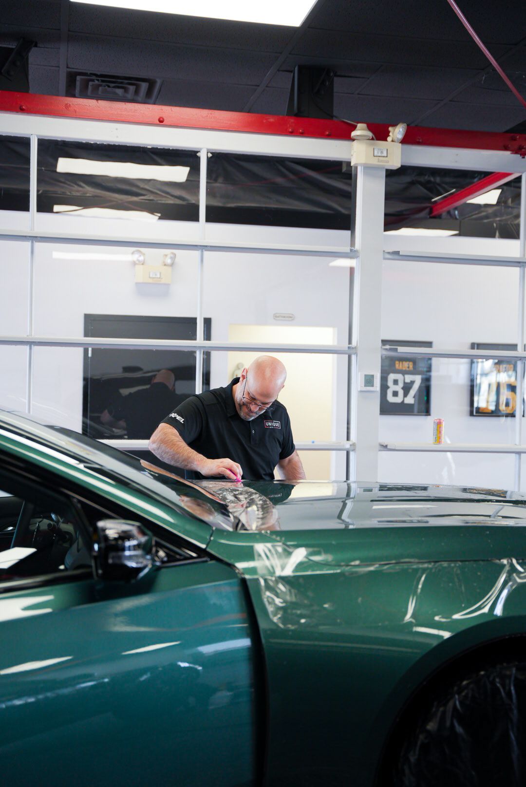 Bald man in a black shirt installing film on a green car in a garage.
