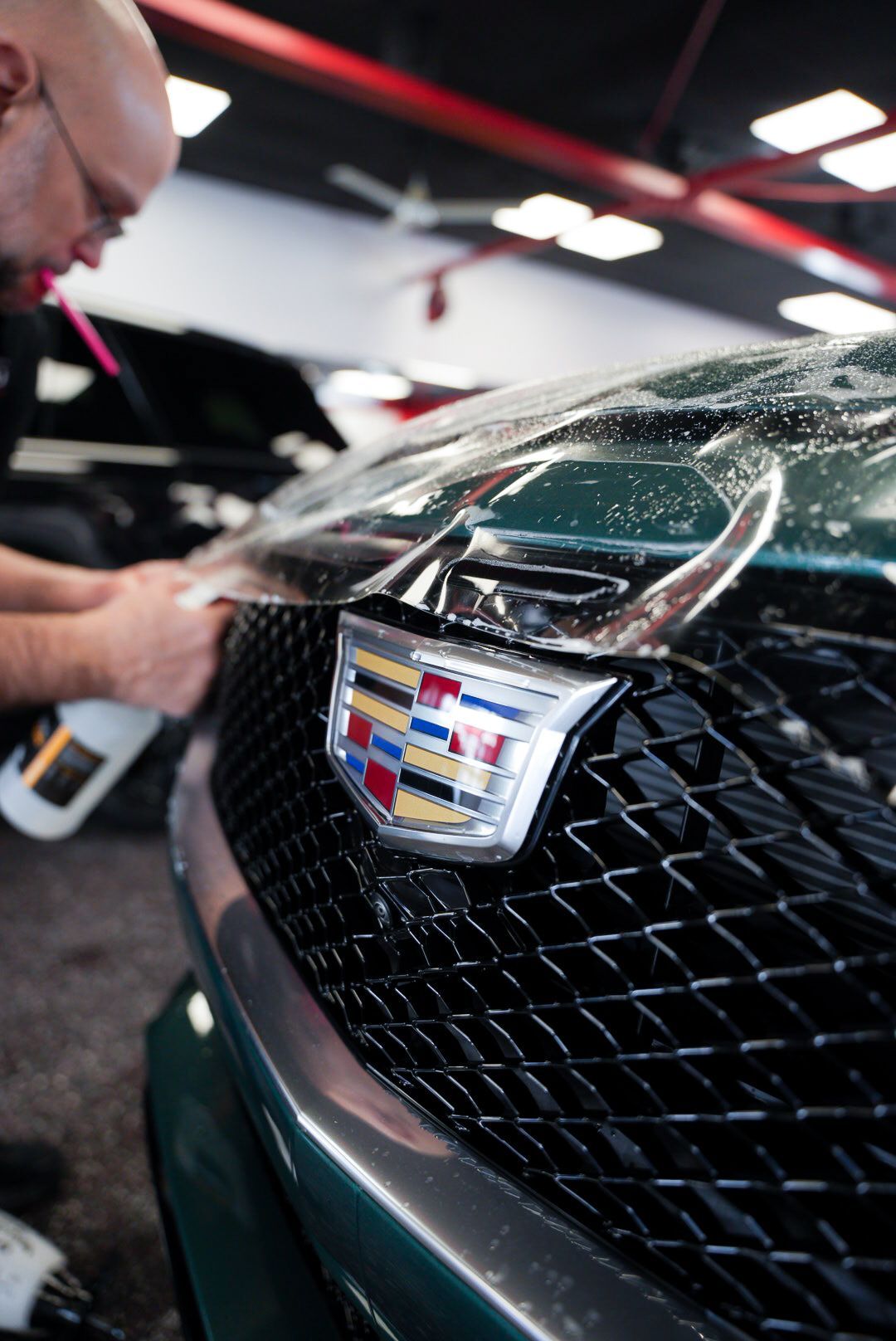 Person applying protective film to the hood of a green Cadillac. Close-up view of the Cadillac emblem and grill.