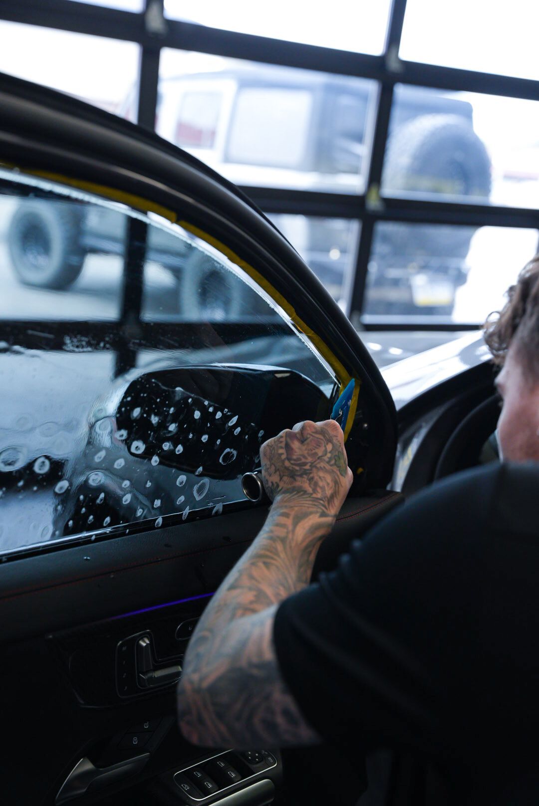 A person with tattoos applying window tint to a car, using a squeegee and water in a garage.