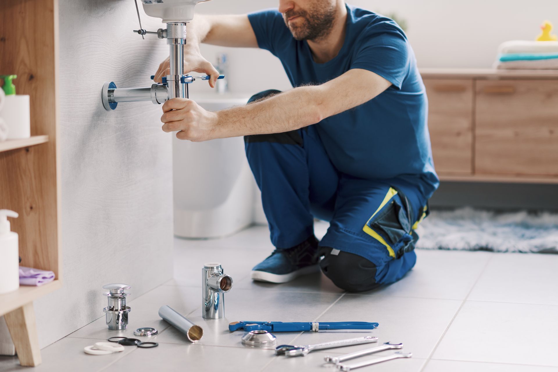 Plumber kneeling, fixing sink pipes in a bathroom. Blue uniform, tools on floor, white walls and sink.