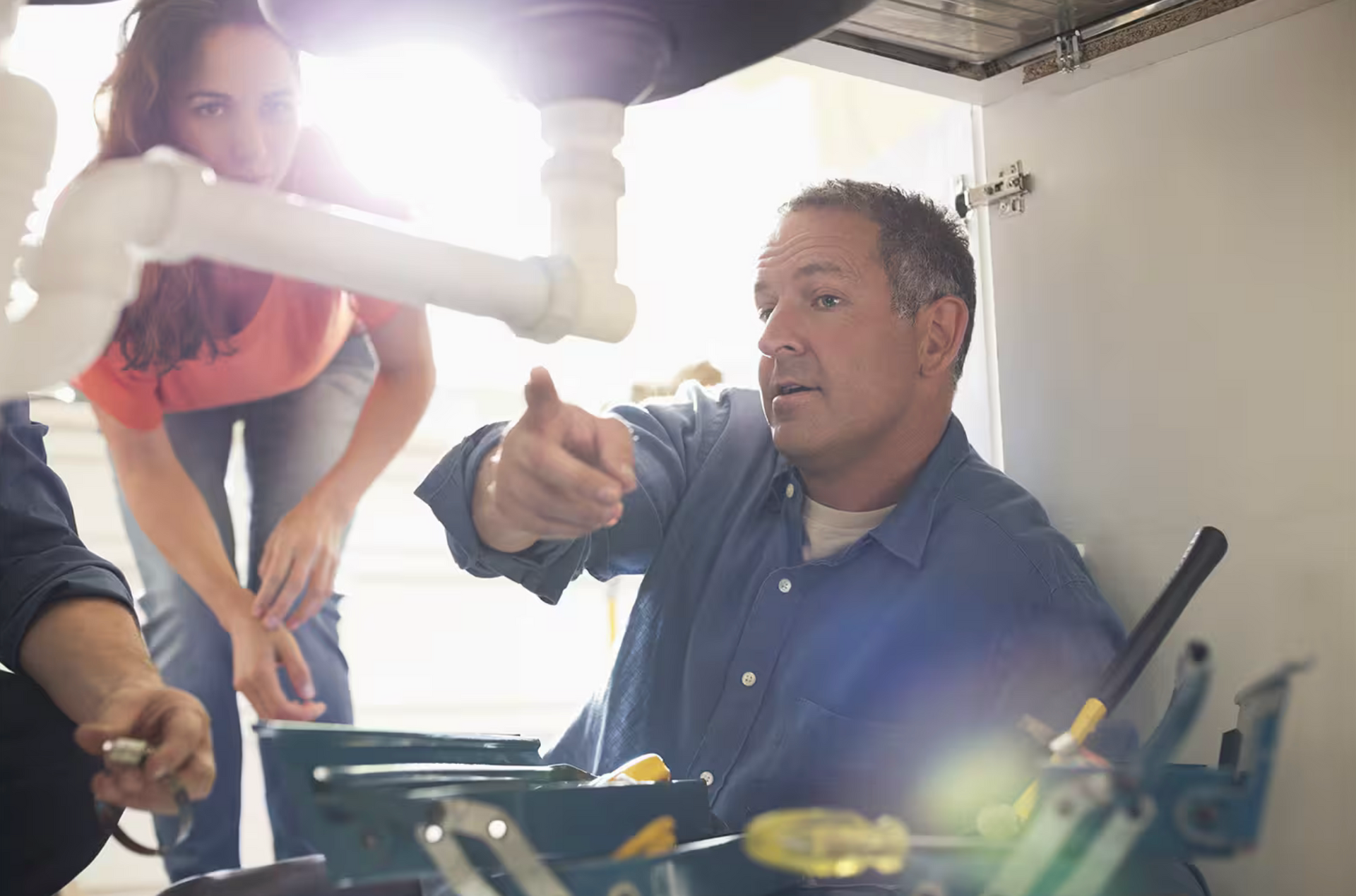 Man pointing at plumbing under a sink as a woman looks on; tools visible in a toolbox.