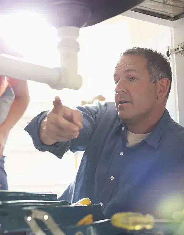 Man pointing at plumbing under a sink as a woman looks on; tools visible in a toolbox.