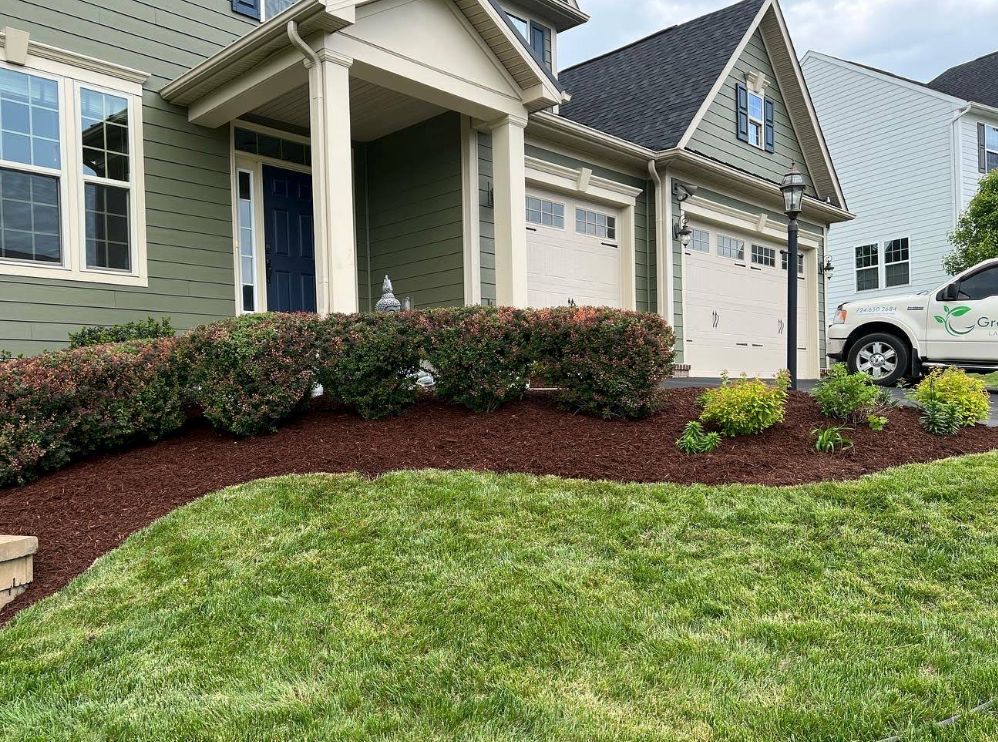 Front yard of a suburban house with shrubs, mulch landscaping, and a garage driveway.