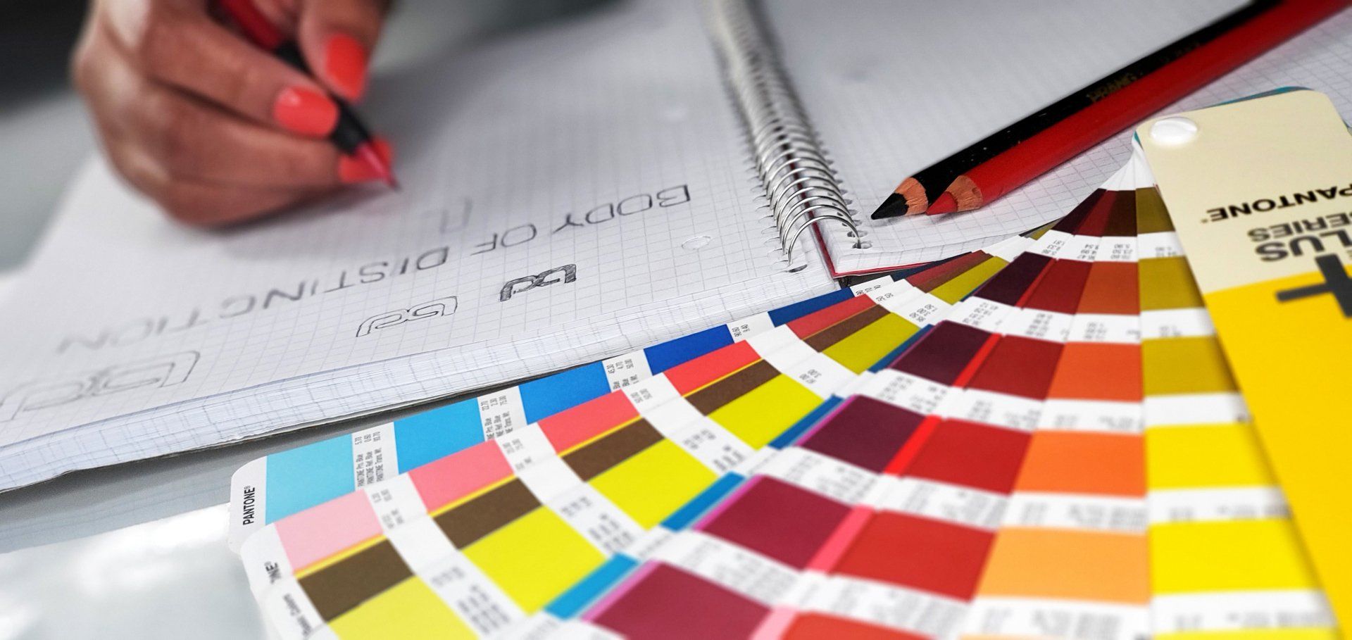 A hand writing in a spiral notebook, with a color swatch and pencil nearby.