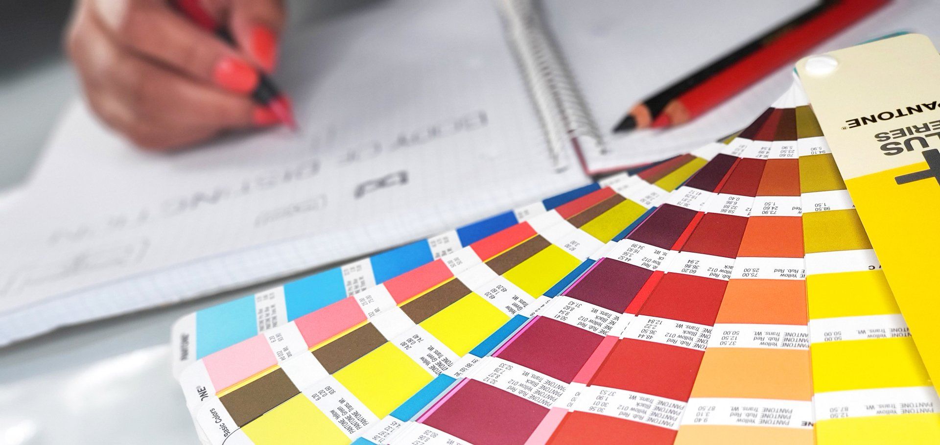 Person writing on a notepad, with a colorful color swatch fan and pencils in the background.