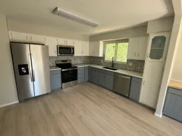 Empty modern kitchen with stainless steel appliances, gray cabinets, granite countertops, and light wood flooring