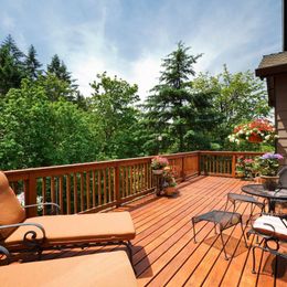 Wooden deck patio with lounge chairs, table, and potted flowers overlooking lush green trees under a blue sky