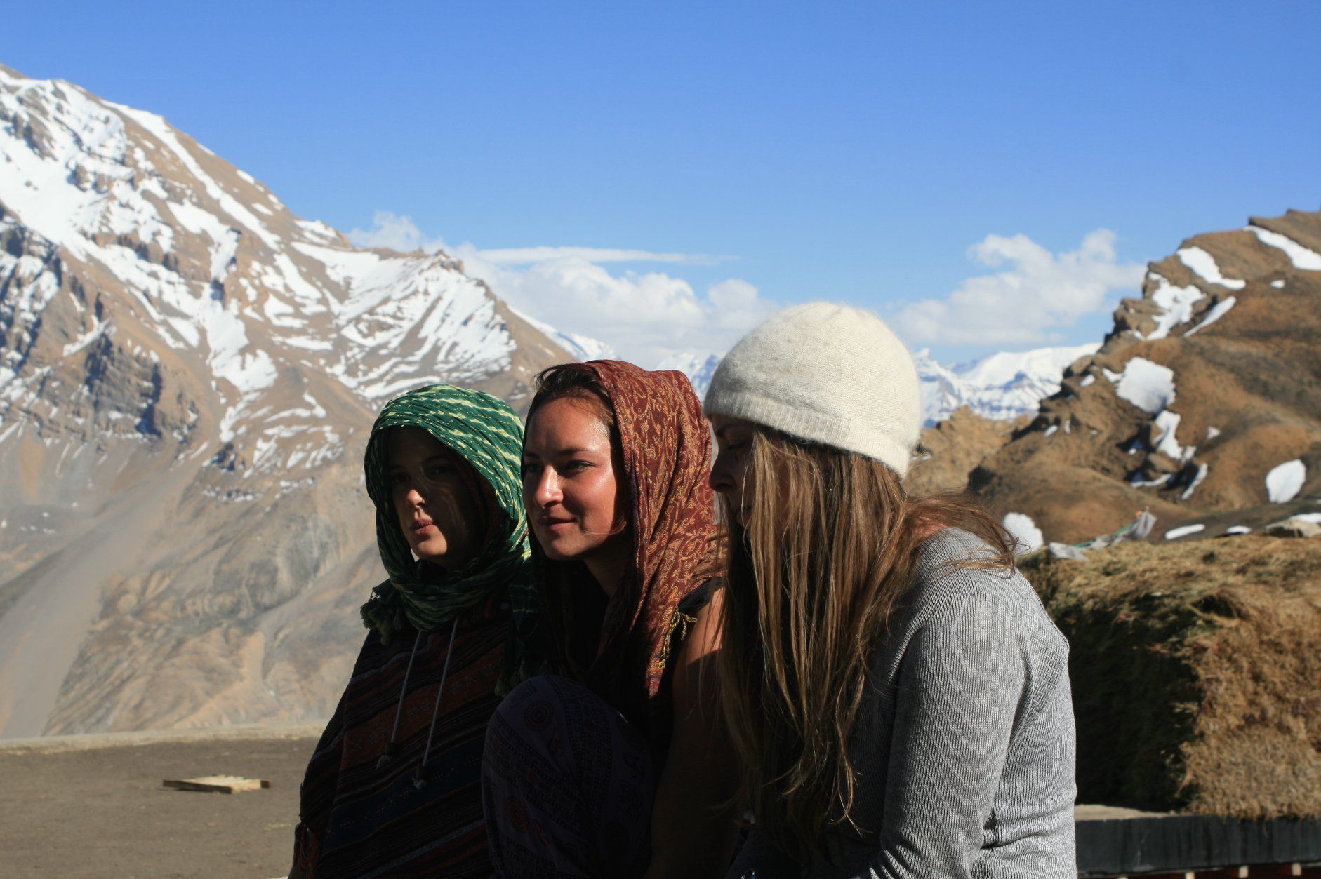 Three people pose outdoors with a snowy mountain backdrop. One wears a green headscarf, another a red one, and the third a white hat.