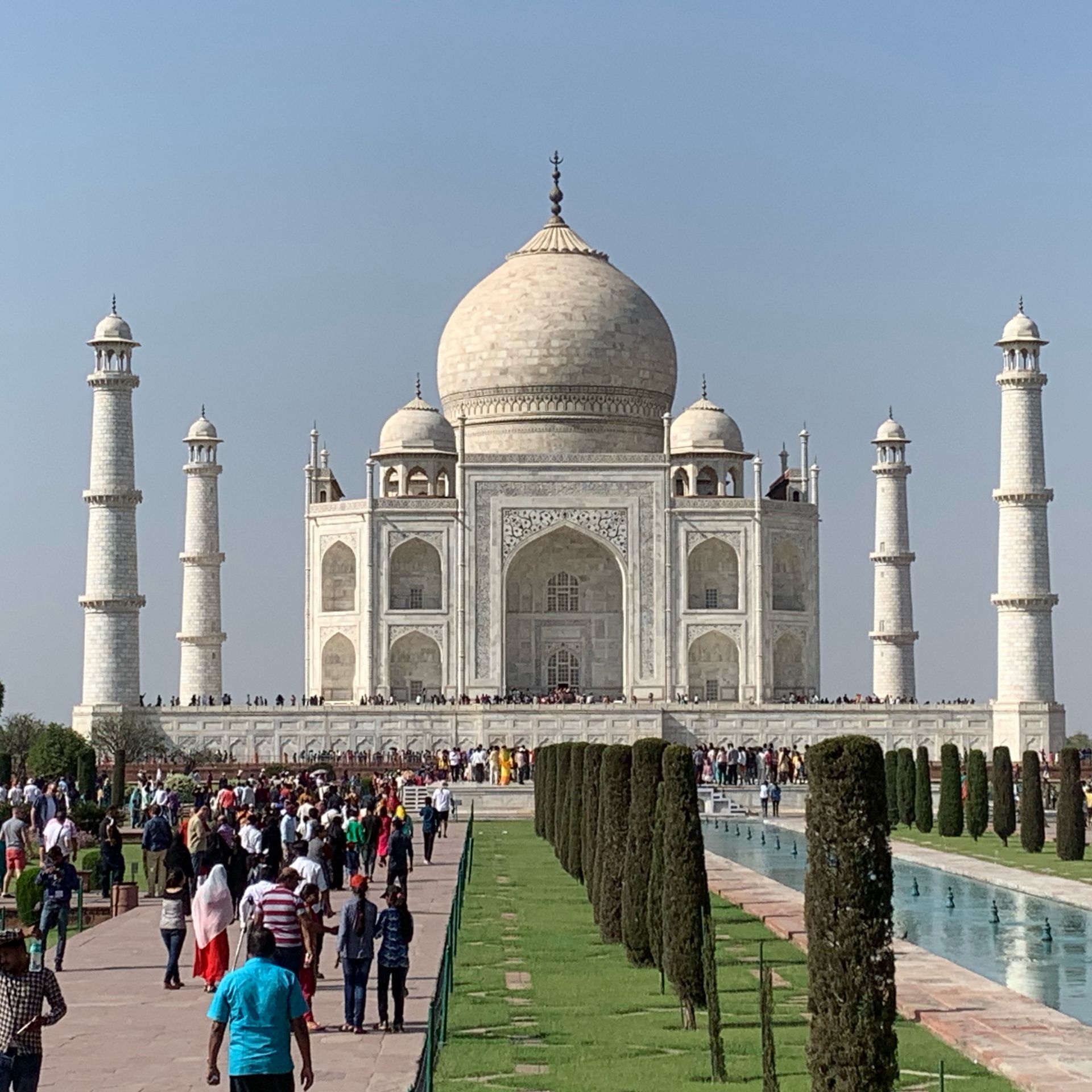 Taj Mahal, white marble mausoleum in Agra, India. People walk on grounds towards the building. Blue sky.