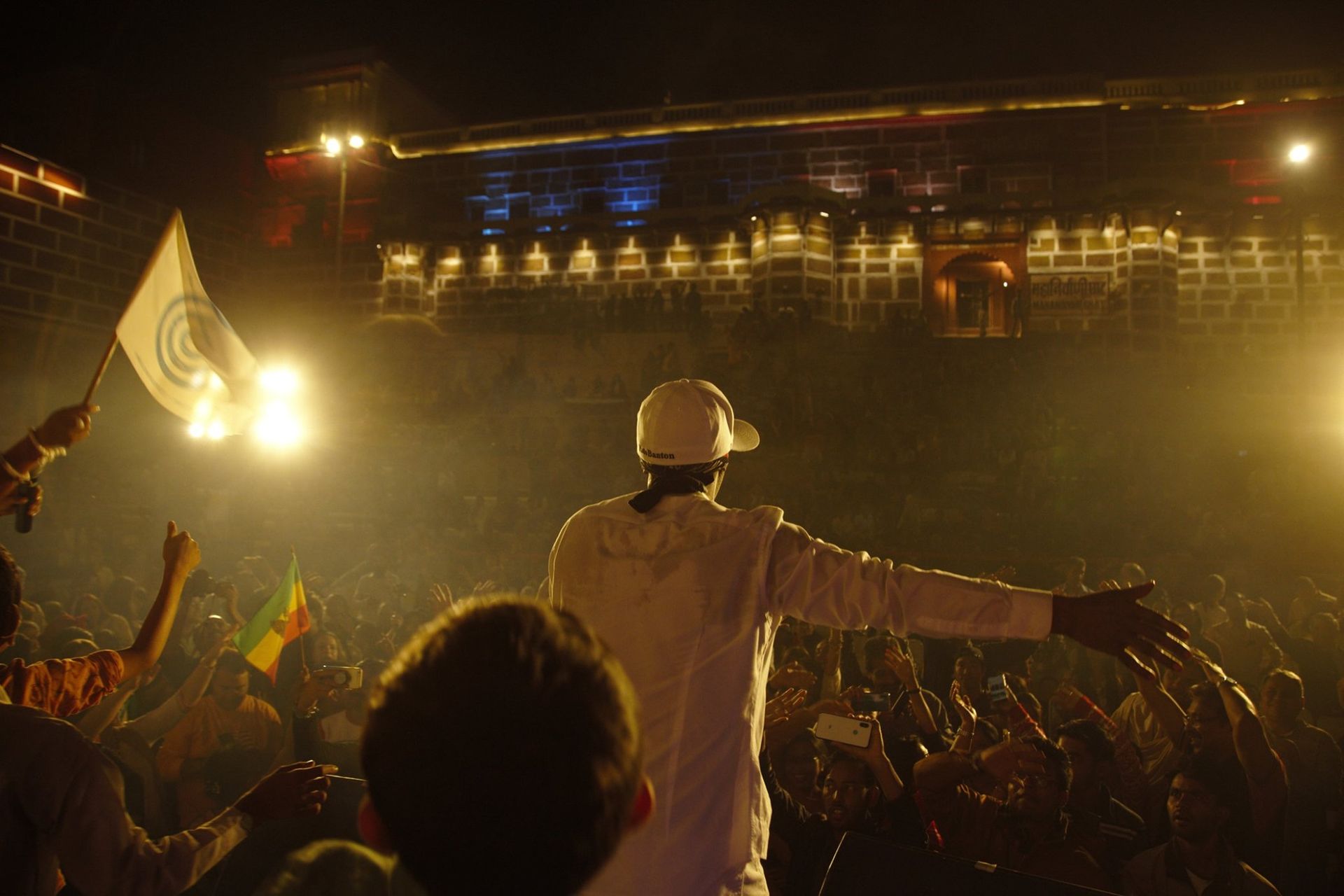 Man in white outfit on stage with outstretched arm, crowd cheering, lit-up building in background at night.
