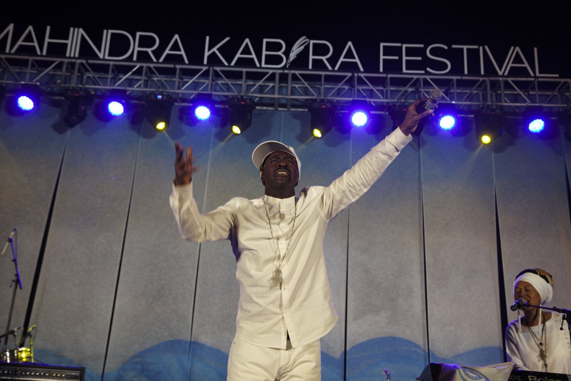Man on stage with arms raised at the Mahindra Kabira Festival. White outfit, blue lights.