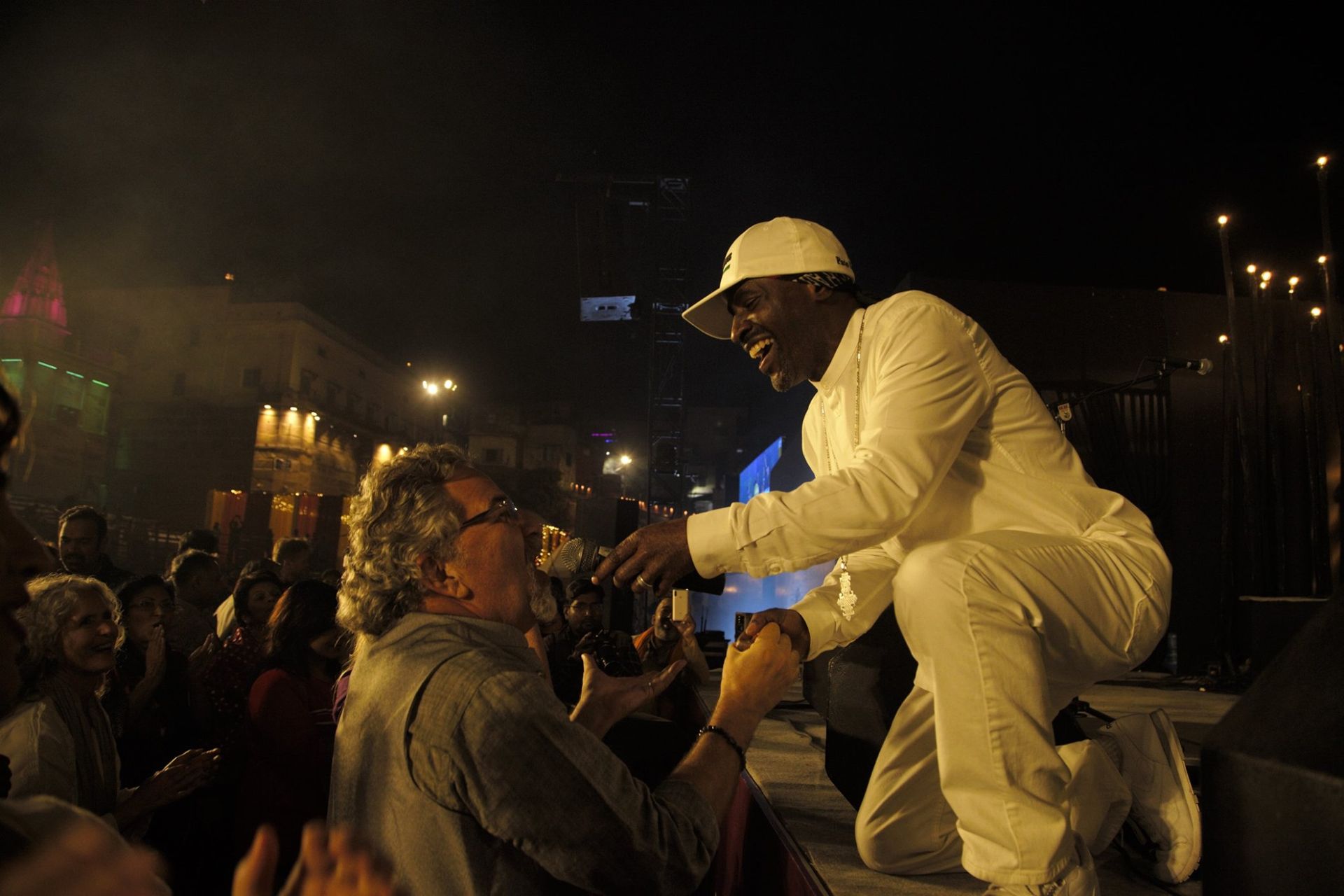 Man in white suit kneels to shake hands with a person in the crowd at an outdoor evening event.