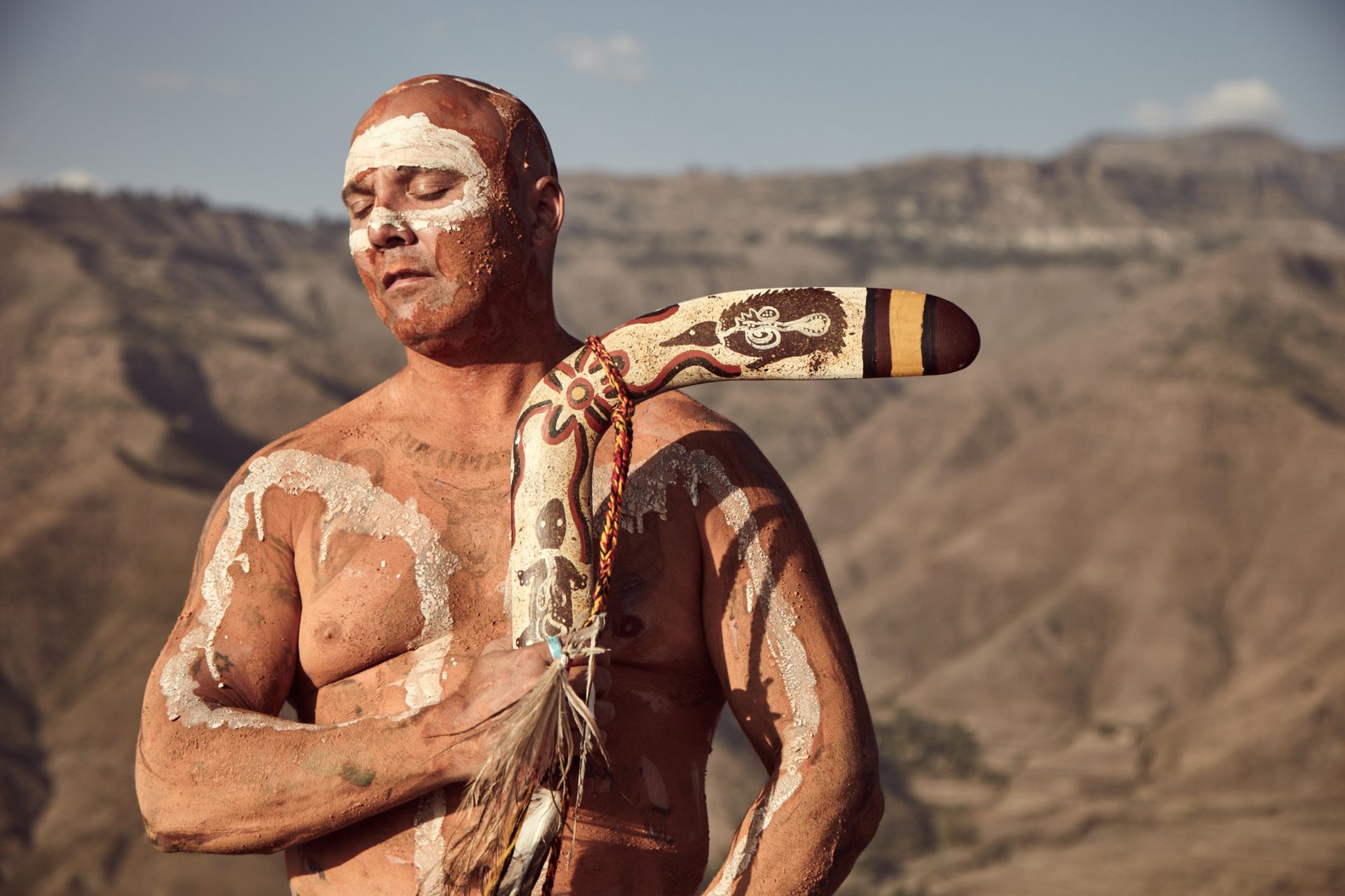 Man with painted body and boomerang, standing outdoors with mountains in the background.