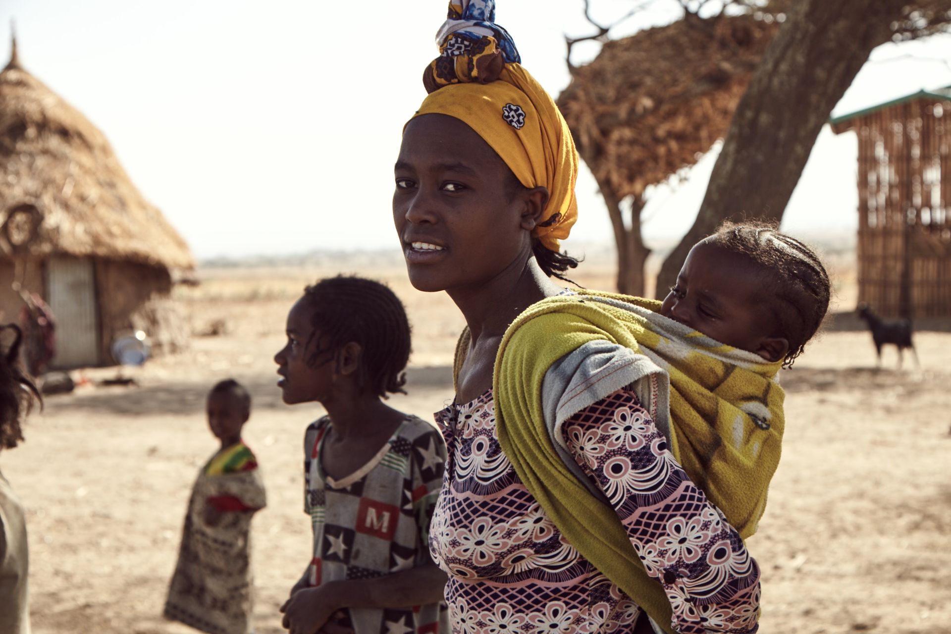 Woman carrying a baby on her back, wearing yellow headscarf, standing near huts in a sunny, dry environment.