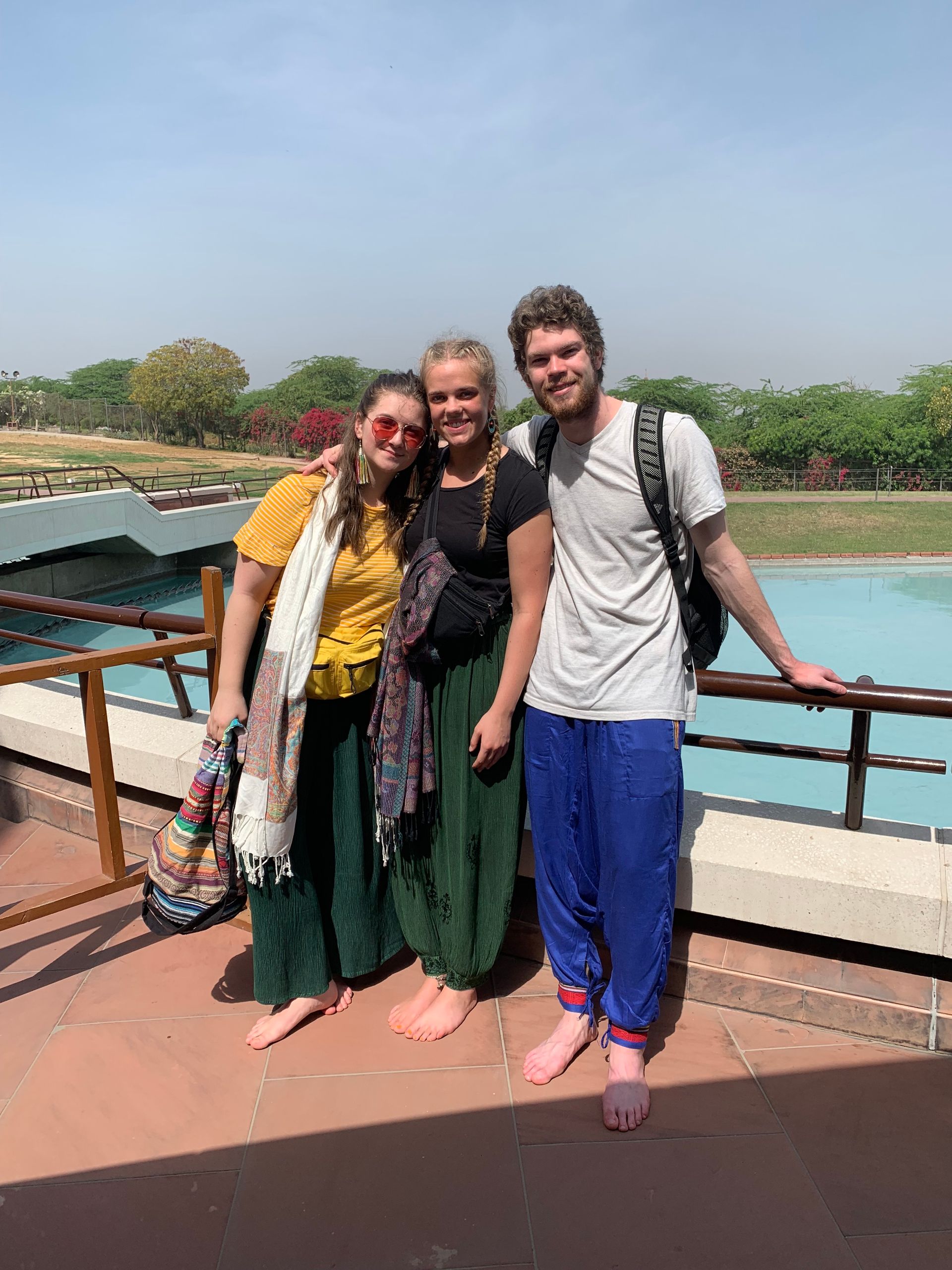 Three people pose on a terrace: one in yellow, one in black, and one in a white shirt and blue pants. A body of water and trees are visible.