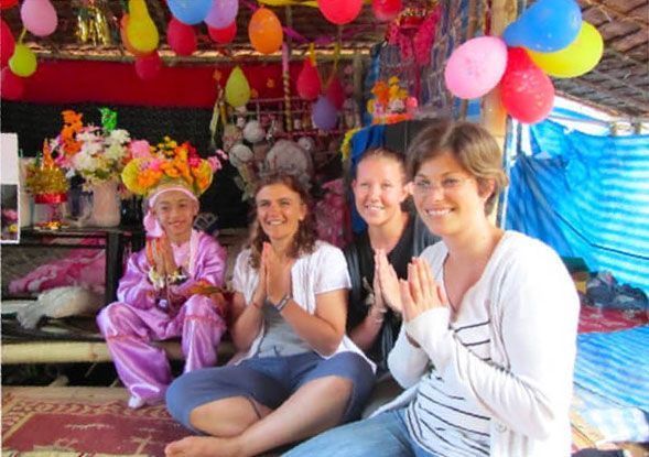 Group of people with hands in prayer pose, smiling, with a person in pink costume sitting nearby, festive decorations overhead.