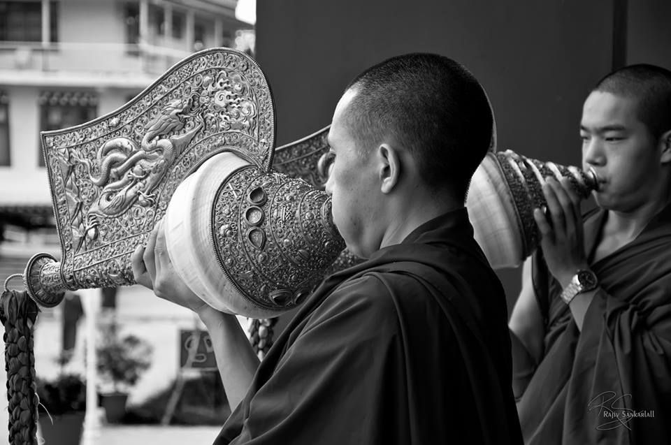 Two monks in brown robes playing large, decorative horns outdoors.