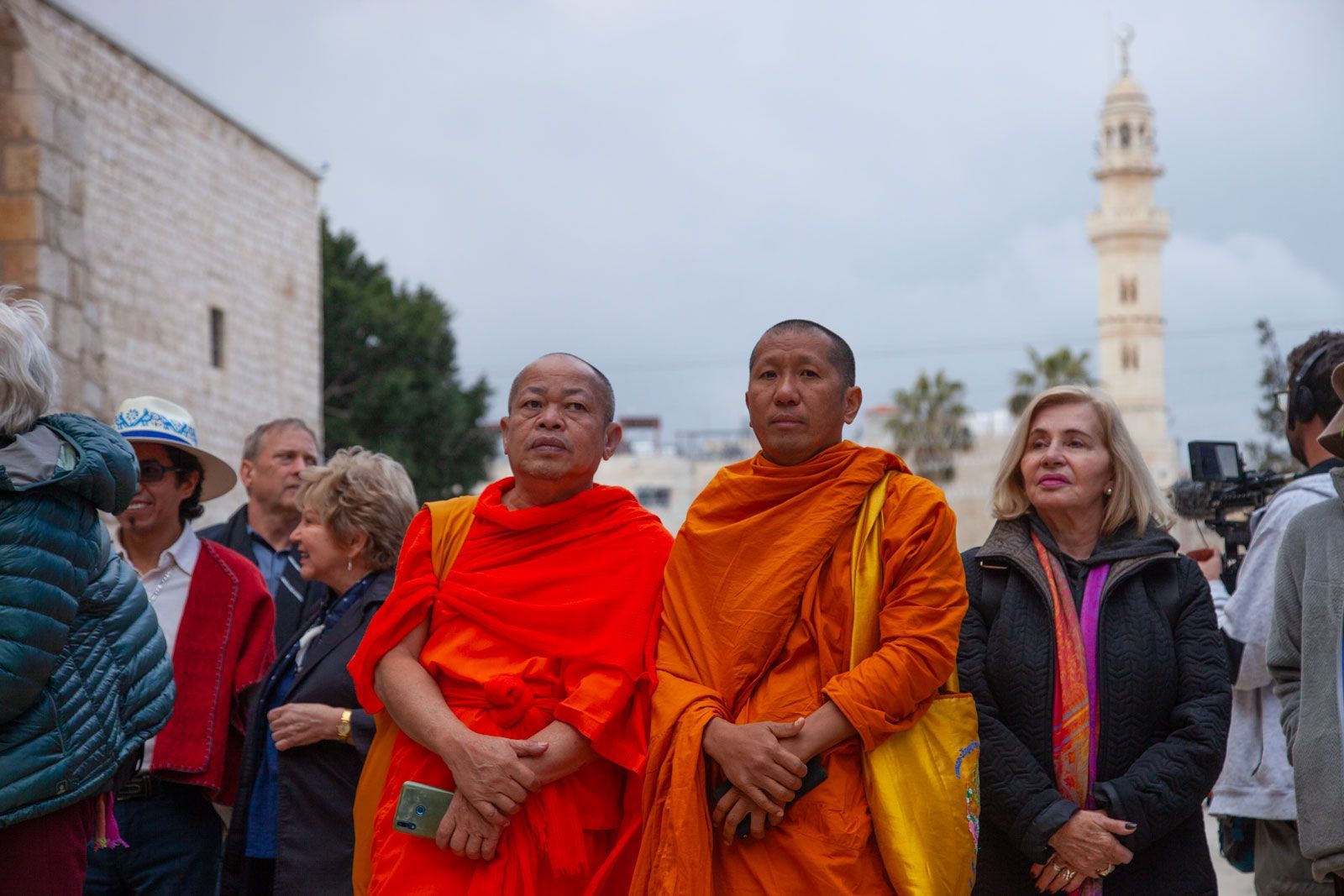 Two monks in orange robes stand outside near a building, others around them.