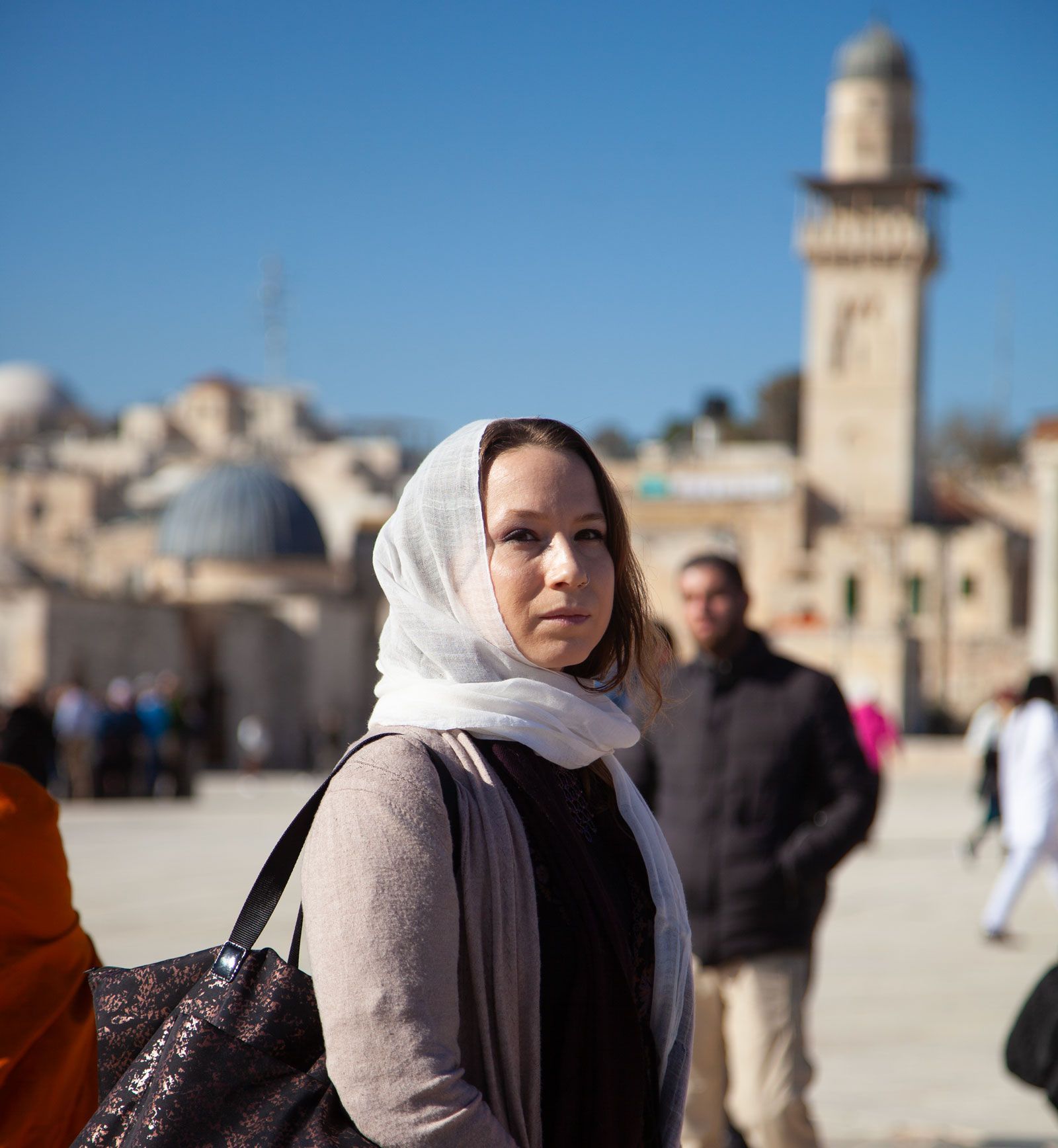 Woman in headscarf, light sweater, and bag, in front of a mosque and people on a sunny day.