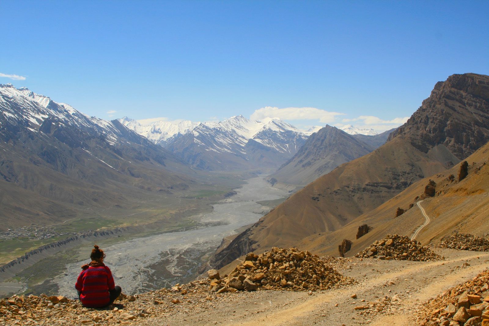 Person in red jacket sits on a hillside, overlooking a valley and snow-capped mountains.