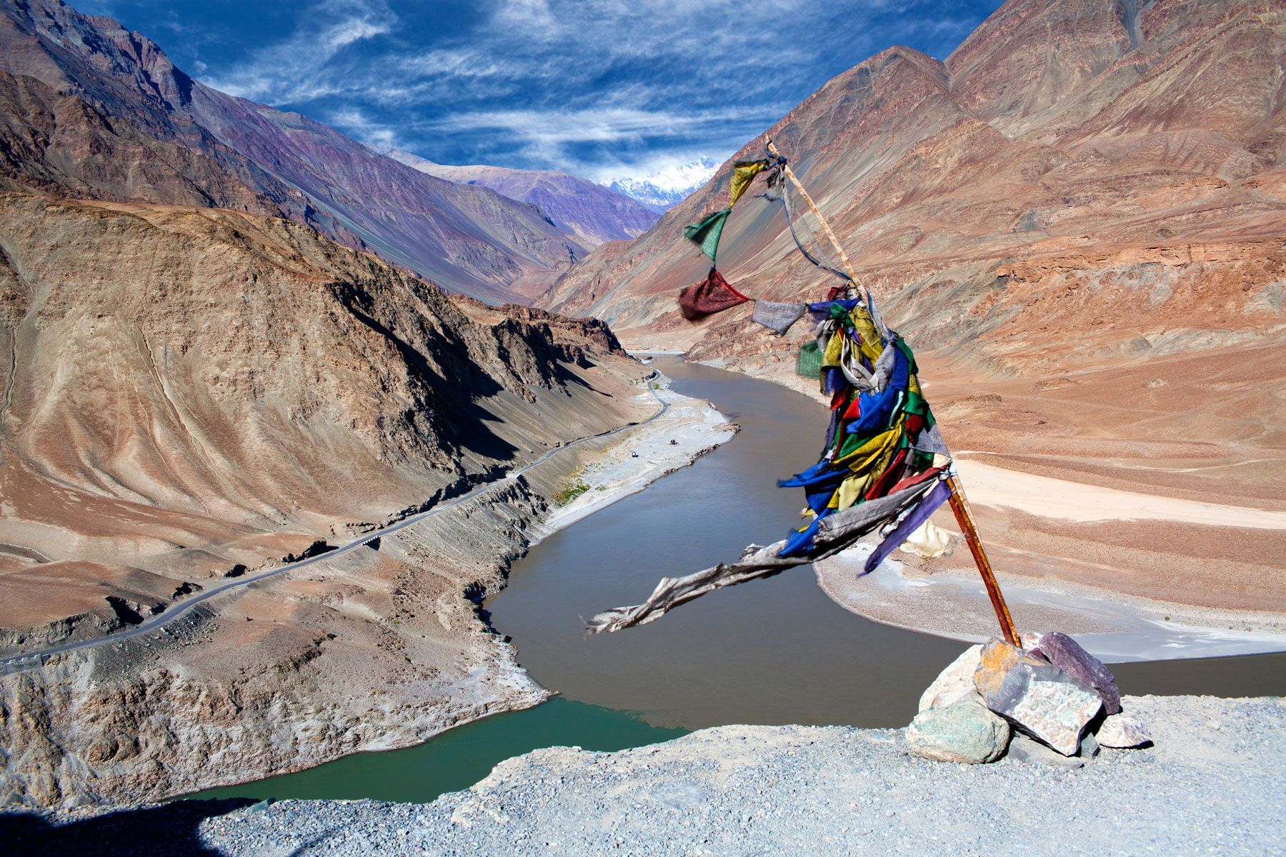 Confluence of Indus and Zanskar rivers in Ladakh, India; brown and green waters meet, mountains in background, prayer flags in foreground.