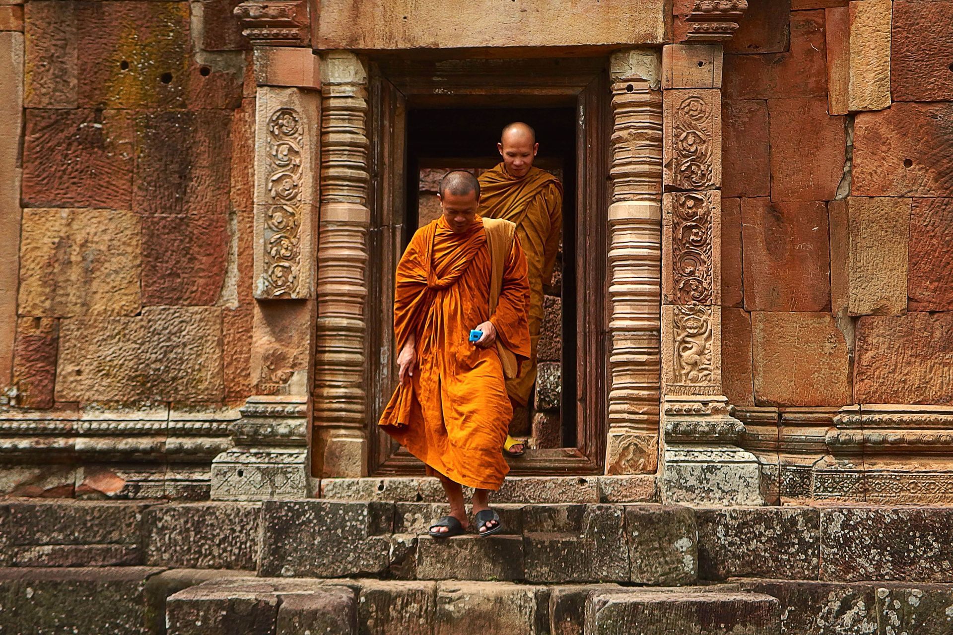Two Buddhist monks in orange robes exit a stone doorway.