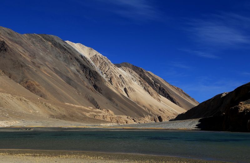 Mountains rising from lake, blue sky. Brown and tan mountains, teal water, clear blue sky.