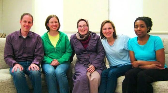 Five people sitting on a beige couch, smiling. Indoors, neutral background.