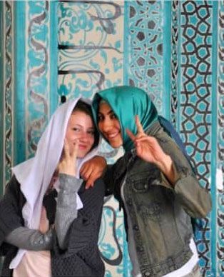 Two people in headscarves pose with peace signs in front of a turquoise tiled wall.