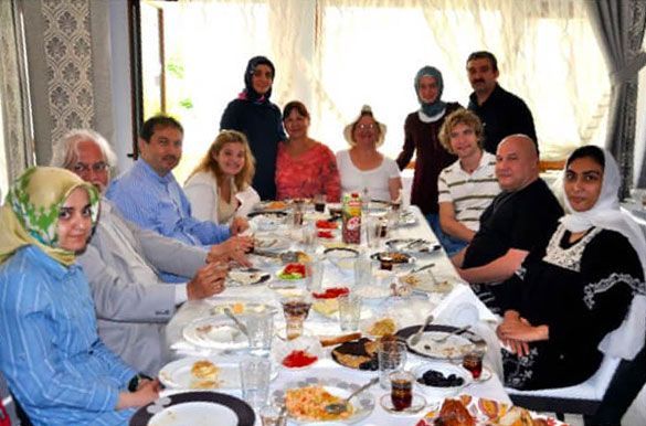 Group of people at a table, eating. Some wear head coverings. Sunlight streams through a nearby window.