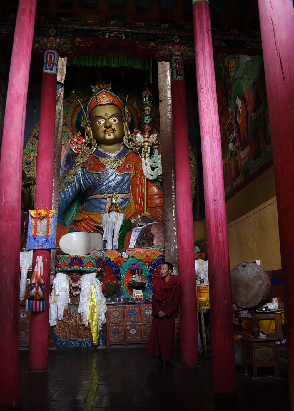 A seated golden Buddha statue inside a temple. A monk stands at the base. Pink pillars frame the space.