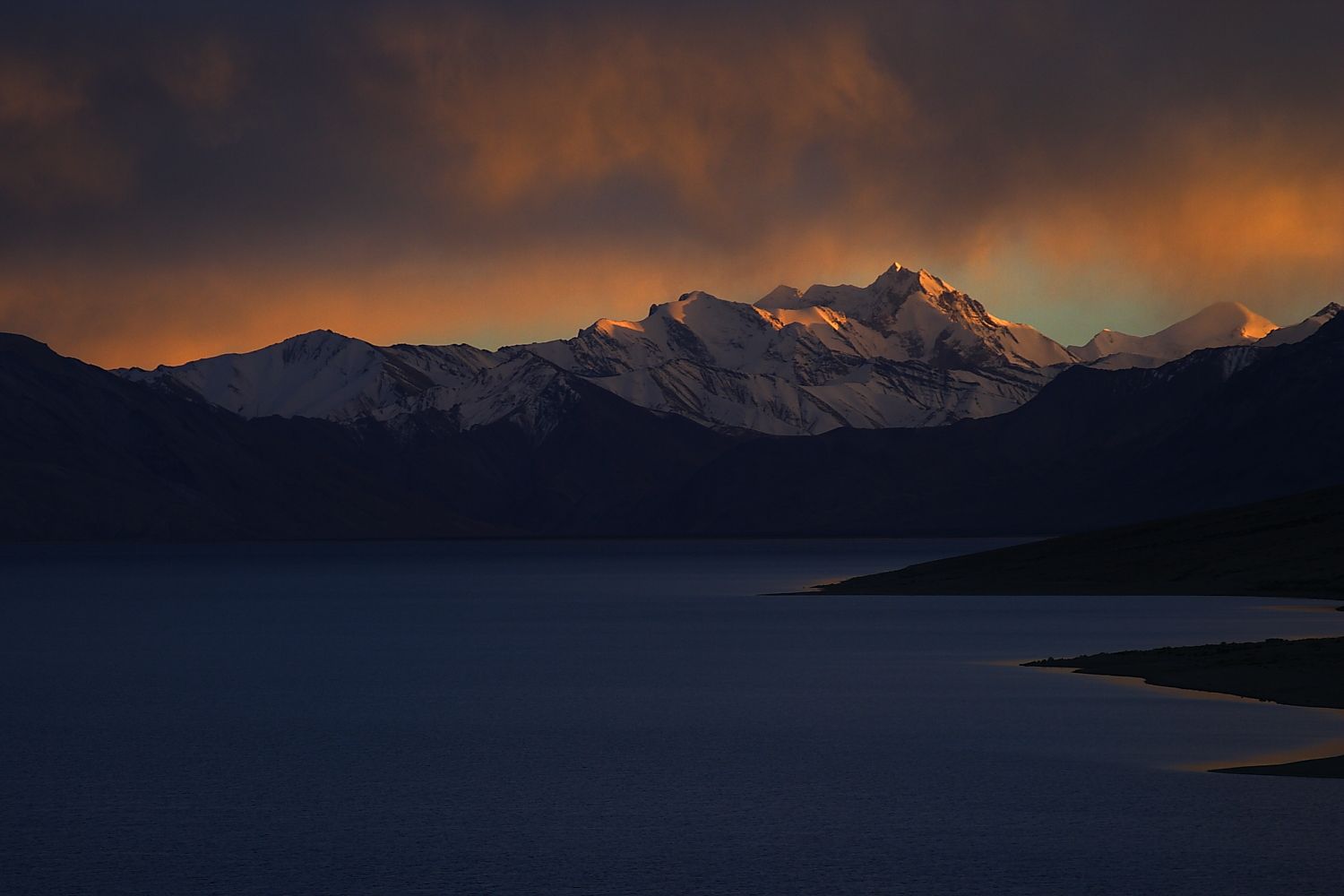 Snow-capped mountains at sunset over a dark lake; golden light reflects on the peaks and clouds.