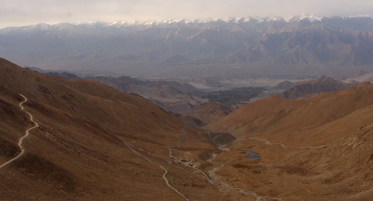 Brown valley winding between arid hills, with distant snow-capped mountains and green vegetation at the bottom.