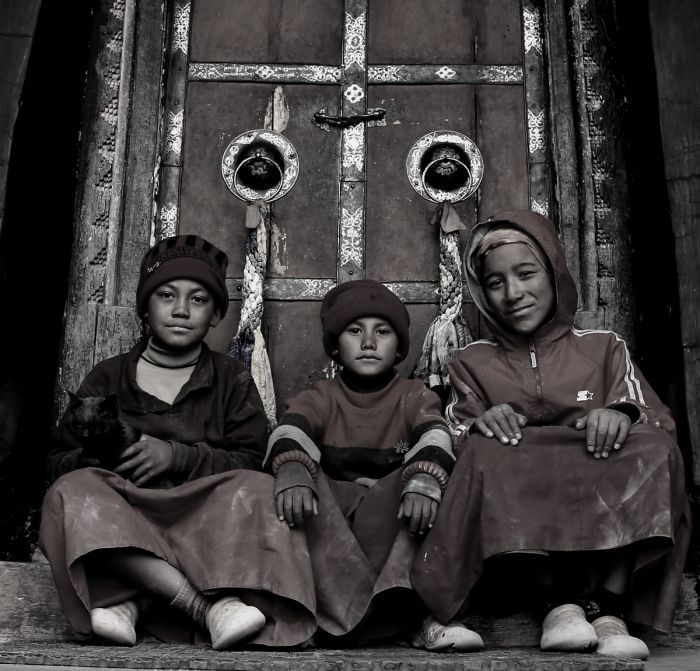 Three children sit in front of a wooden door. They wear head coverings and robes, looking at the camera. Black and white photo.