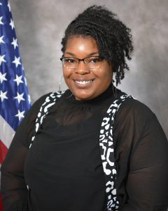 A woman wearing glasses is standing in front of an american flag.
