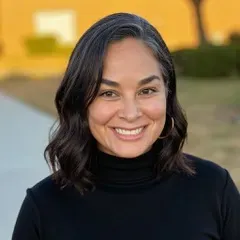 A woman with long brown hair is smiling and wearing a white shirt.