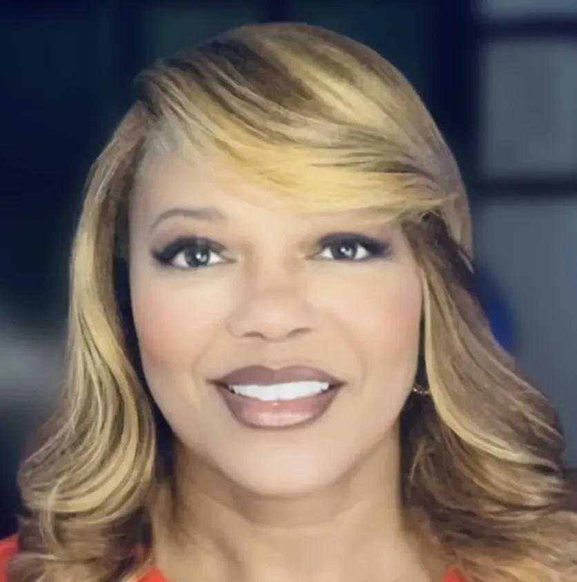 A woman in an orange shirt and pearl necklace is smiling for the camera.