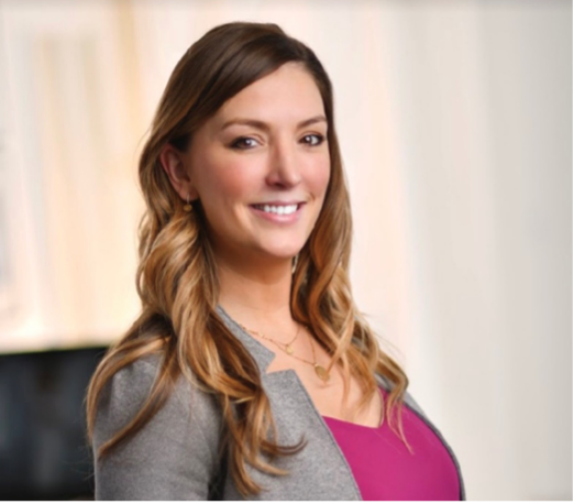 A woman in a pink shirt and grey jacket smiles for the camera