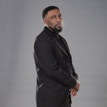 A man in a black suit and tie is standing in front of a gray background.