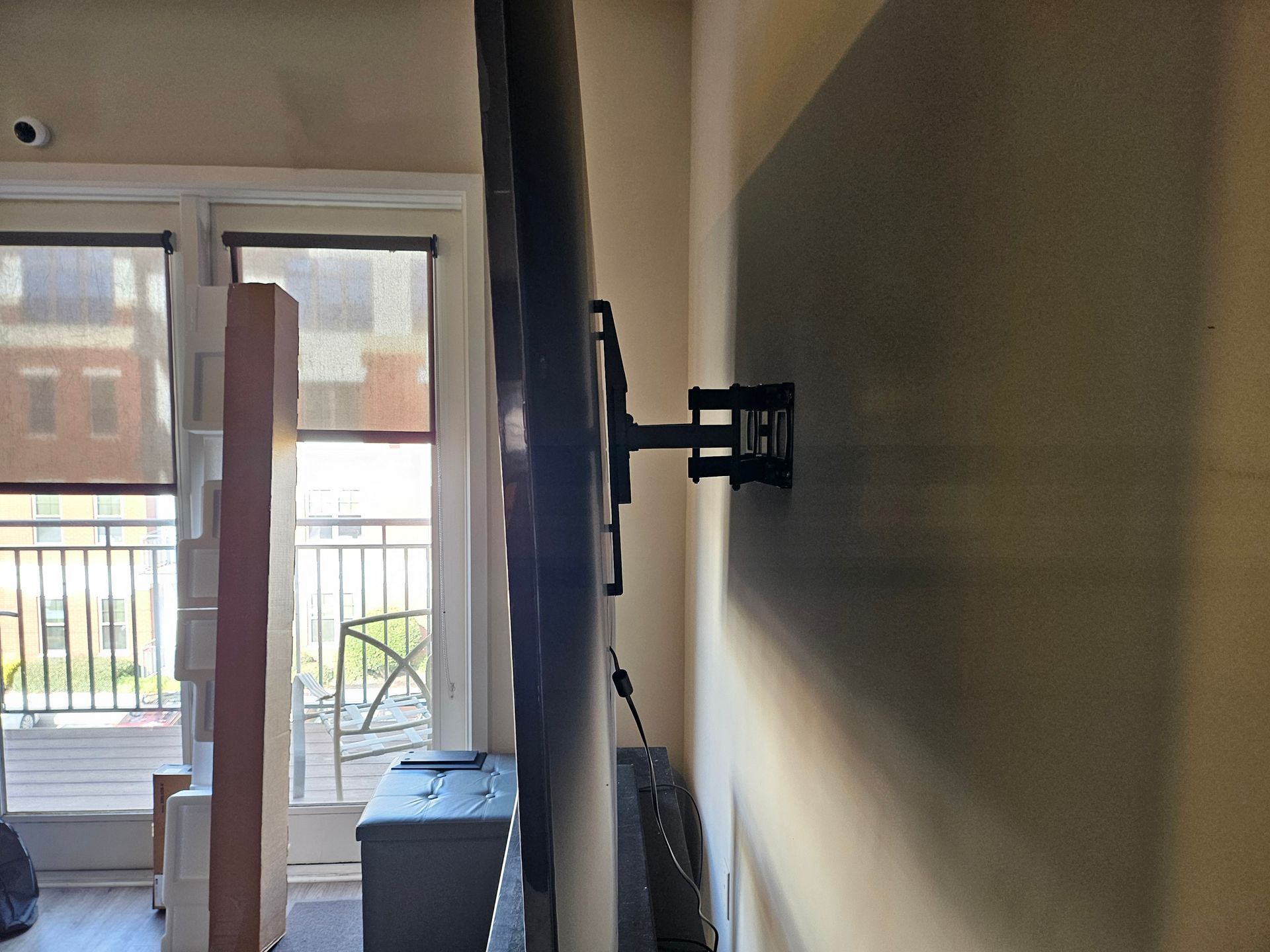 TV mounted on a wall with a black articulating arm. The wall is tan, with a window and balcony in the background.