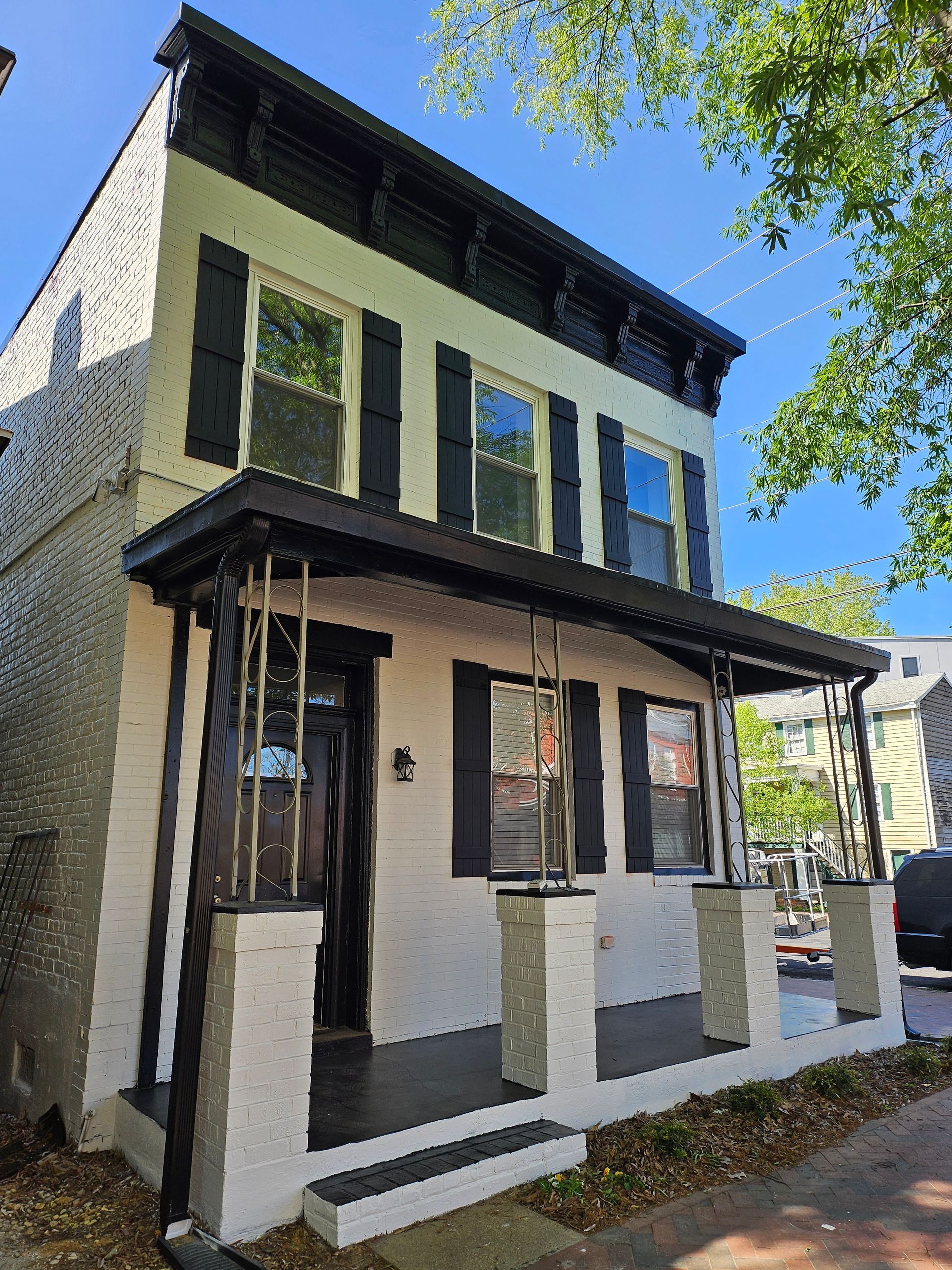 Two-story white brick building with black trim and shutters, featuring a porch supported by pillars.